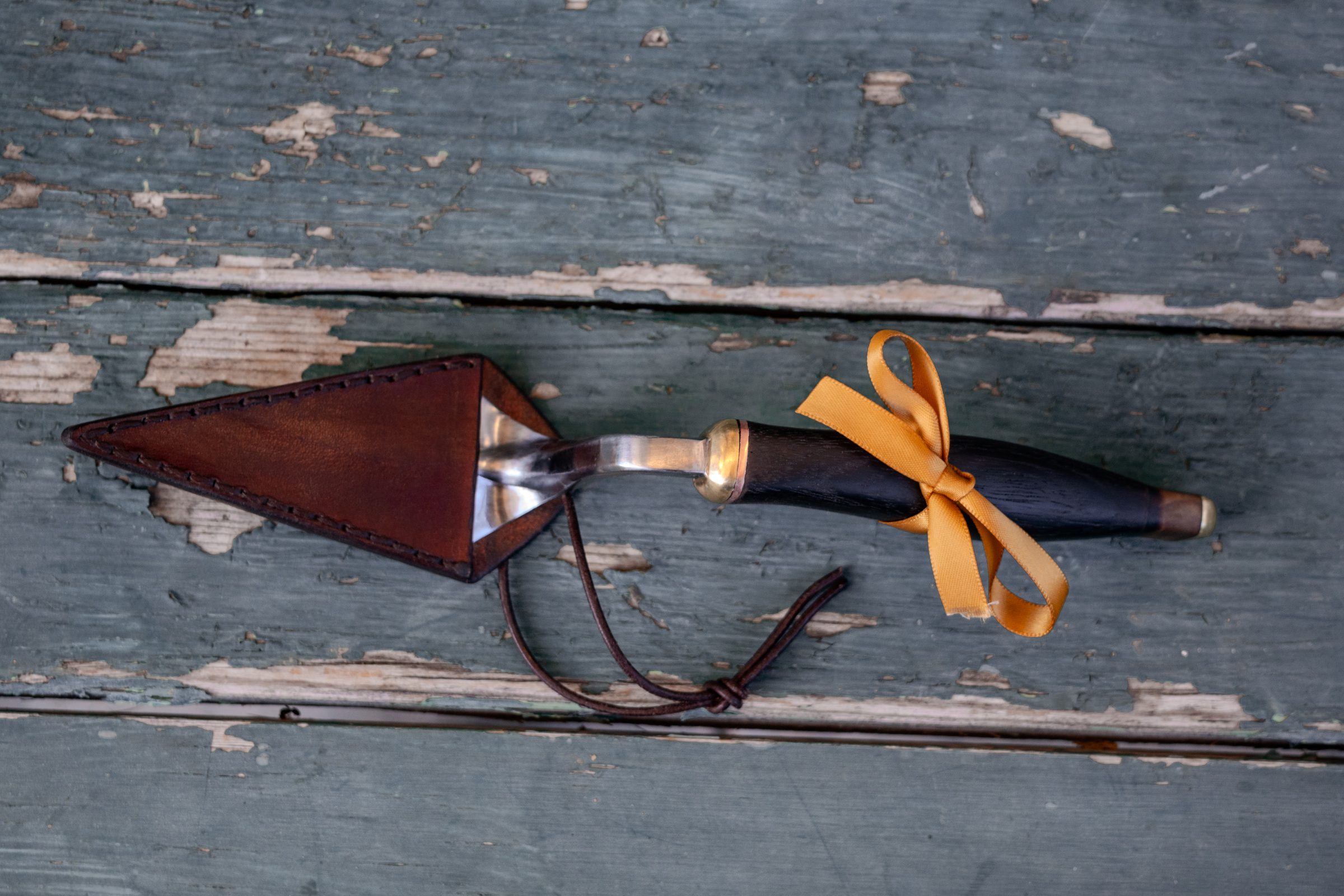 Hand-forged archaeologist's trowel with its leather sheath, resting on weathered painted wood